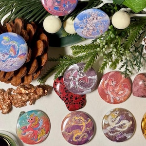 May include: A set of nine colorful round buttons featuring various dragon and unicorn illustrations. The buttons are arranged on a white surface with a few decorative items, including a pine cone, a cinnamon stick, and a gold ornament.