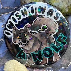 May include: A circular badge featuring a cartoon wolf design. The badge has the text "OMISHOWAS" in white and "AWOLF" in teal. The wolf is grey and brown with yellow eyes, set against a dark, glittery background.