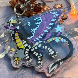 Puede incluir: Un llavero acrílico transparente con un diseño de dragón con detalles en negro, morado, amarillo y blanco. El dragón tiene alas grandes y detalladas y una larga cola. El llavero incluye un cierre en forma de estrella.
