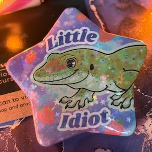 Puede incluir: Pin en forma de estrella con una ilustración de un gecko de dibujos animados. El pin presenta las palabras "Little Idiot". El gecko es verde y sonríe, sobre un fondo de tonos rosas y azules con detalles de purpurina.