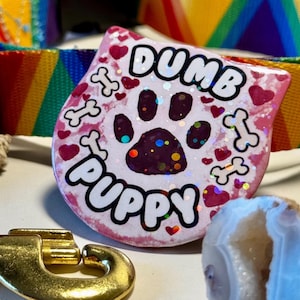 May include: Pink dog tag with the text "Dumb Puppy" in white lettering, surrounded by pink hearts and white bone shapes. A sparkly paw print is in the center of the tag.
