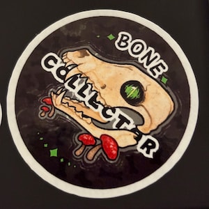 May include: A round sticker with a black background featuring a skull with the text "BONE COLLECTOR" in white. The skull is tan with a green eye and red mushrooms.