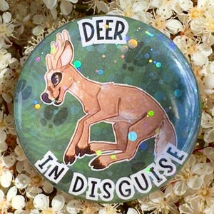 May include: A round button with a cartoon deer illustration. The deer is brown with blue accents, set against a green background with the words "DEER IN DISGUISE". The button has a glittery, iridescent effect.