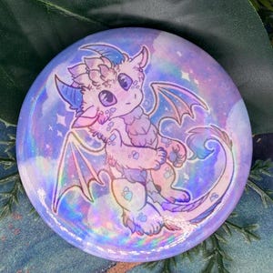 Puede incluir: Un botón redondo con un acabado holográfico presenta una ilustración de dibujos animados de un dragón morado y rosa con alas y cola. El dragón está sobre un fondo de nubes moradas y azules que se arremolinan.