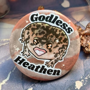 May include: A round button with a cartoon gecko illustration. The gecko has brown spots and is set against a pink and gray background. The words "Godless Heathen" are printed in black, curved around the gecko.