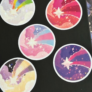 Puede incluir: Cinco pegatinas redondas con un diseño de estrella y estrella fugaz colorida. Cada pegatina presenta una combinación de colores diferente, incluyendo arcoíris, rosa, rojo, morado y amarillo. Las pegatinas tienen un borde blanco sobre una superficie negra.