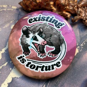 May include: A round button with a distressed pink and brown background. The button features a crying opossum illustration with the words "existing is torture" in white text. The opossum is gray, black, and pink.