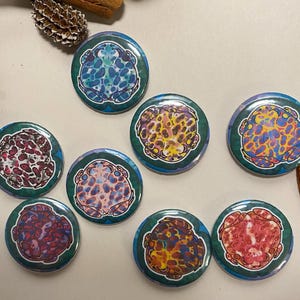 May include: Eight round buttons with a green background and a colourful frog design. Each frog has a different colour pattern, including red, yellow, blue, and pink.