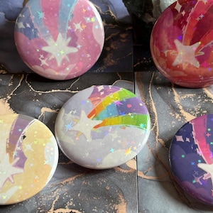 May include: A selection of circular pin badges with a star and shooting star motif. The badges display a range of colours, such as pink, purple, and blue, with iridescent highlights. The badges are arranged on a patterned surface.