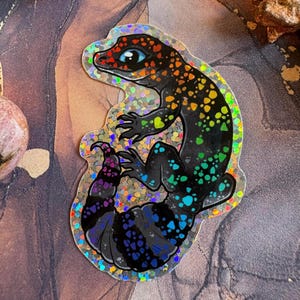 May include: A holographic sticker of a lizard with a rainbow design. The lizard has a black body with colourful spots and a red head. The sticker has a glittery, iridescent border.