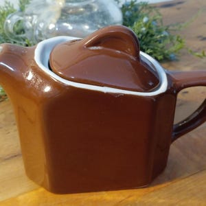 May include: A brown ceramic teapot with a lid, white trim, and a handle. The teapot has a square shape and a spout. The background includes a glass teapot and greenery.