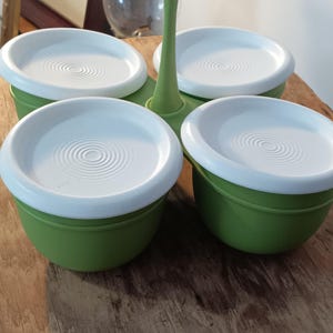May include: A green and white serving dish set. The set includes four round, green bowls with white lids. The lids have a concentric circle design. The bowls are connected by a green handle.