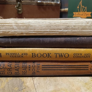 May include: A stack of four vintage books with aged covers. The top book has a light tan, textured cover. The second book has a dark brown cover. The third book is yellow with the text "BOOK TWO". The bottom book is brown with the text "BOOK FIVE".