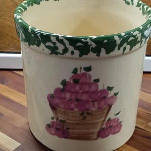 May include: Cream-colored ceramic utensil holder with a green and white rim. The holder features a hand-painted design of a basket filled with pink apples. The holder has a crackle finish.