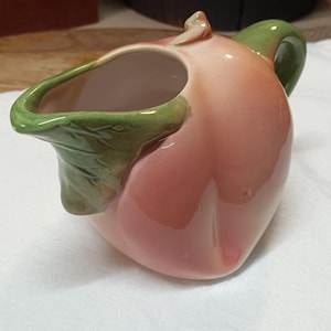 May include: A ceramic pitcher shaped like a peach, with a pink body and green handle and spout. The pitcher has a glossy finish and is set against a white background.