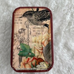May include: A small, rectangular tin box with a vintage design. The box features a black bird with a white breast, a fairy with wings, and a rose with a butterfly. The text on the box reads "at the picture Fair".