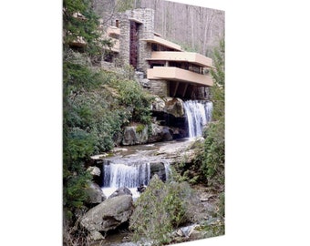 May include: A modern, beige-colored house with a flat roof and large windows sits on a rocky cliff overlooking a cascading waterfall. Lush green trees and foliage surround the house and waterfall.