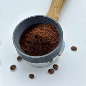 Könnte beinhalten: Ein grauer Siebträger mit einem Holzgriff, gefüllt mit dunkelbraunem Kaffeepulver. Der Siebträger ist von mehreren ganzen Kaffeebohnen auf einer weißen Oberfläche umgeben.