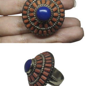 May include: A silver ring with a large, round face. The face features a central blue stone surrounded by coral-colored rectangular stones and silver accents. The ring is shown from two angles.