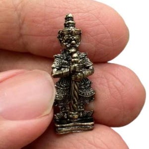 Pode incluir: Pequena e detalhada estatueta de bronze de um guerreiro em pé, segurando um bastão. A figura é adornada com uma armadura intrincada e uma coroa. A estátua é segurada entre dois dedos sobre um fundo branco.