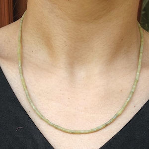May include: A fine necklace made of small, pale green beads. The beads are threaded together to create a thin strand, set against a black top. The necklace has a handmade quality, with a natural, earthy appearance.
