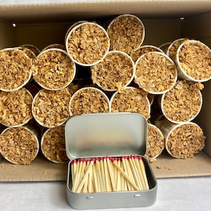 May include: A cardboard box filled with small, white paper cups packed with brown material. A small, open, silver tin of wooden matches with red tips sits in front of the cups. The cups are arranged in rows.