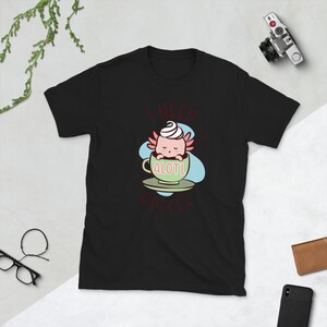 Könnte beinhalten: Schwarzes T-Shirt mit einem Aufdruck eines Axolotls in einer Kaffeetasse. Der Axolotl trägt einen rosa-weißen Wirbel auf dem Kopf und der Text "Ich brauche viel Axolotl-Kaffee" ist auf dem T-Shirt gedruckt.