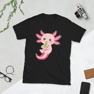 May include: Black t-shirt with a pink axolotl cartoon illustration eating ramen noodles. The axolotl is holding a bowl of ramen with chopsticks and has a happy expression.