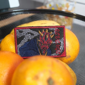 May include: A rectangular patch featuring a red-faced character with horns, a blue cloak, and the word "REVENGE" in white script. The patch is set against a black background with a red border. The patch is placed on top of oranges and lemons in a black wire basket.