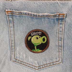 Peut inclure: Une poche en jean bleu avec un patch brodé noir et vert représentant une plante de dessin animé avec le texte "SHOT!"