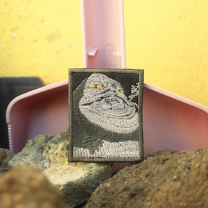 Peut inclure: Écusson brodé représentant une créature grise stylisée aux yeux jaunes sur fond noir, bordé d'un cadre vert foncé. L'image est posée sur un fond jaune avec une pelle rose et des rochers.