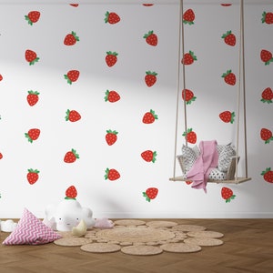 Könnte beinhalten: Eine weiße Wand mit einem sich wiederholenden Muster aus roten Erdbeeren. Ein Holzstuhl mit einem sternförmigen Kissen steht vor der Wand. Eine rosa-weiße Decke hängt an einer Holzschaukel.
