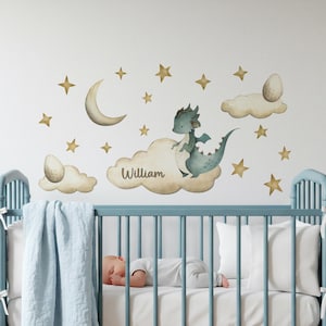 May include: A nursery wall decal set featuring a watercolor dragon, clouds, stars, and a crescent moon. The name "William" is written on a cloud. The scene is set above a blue crib with a sleeping baby.
