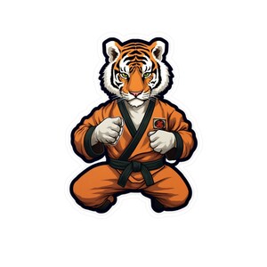 Puede incluir: Una pegatina con un tigre de dibujos animados con uniforme de karate. El tigre es naranja y blanco con rayas negras, lleva un gi naranja con cinturón negro. El tigre está en posición de combate con los puños cerrados.