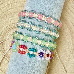 Könnte beinhalten: Vier handgefertigte Armbänder mit Blumenmustern aus runden Perlen und kleinen Saatperlen. Die Armbänder bestehen aus hellem Kordelmaterial und enthalten rosa, weiße, hellgrüne, rote, blaue und violette Perlen, die in Blumenformen angeordnet sind.