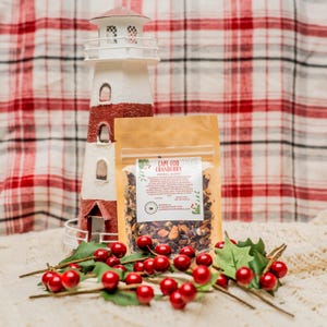 May include: A small brown pouch with a clear window, containing a blend of dried herbs and fruit. The pouch is labeled "Cape Cod Cranberry Herbal Blend". The pouch is sitting on a white surface with red berries and green leaves.