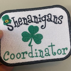 May include: Embroidered patch with a white background, black border, and the words "shenanigans coordinator" in green script. A green shamrock and leprechaun hat are also featured. The patch is rectangular with rounded corners.