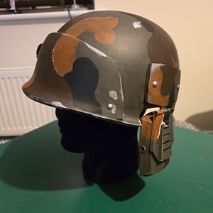 Può includere: Un casco mimetico con accenti marroni e verdi. Il casco ha un pannello laterale con un design testurizzato e una piccola patta.