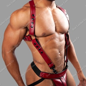 May include: A red leather harness with black hardware. The harness features adjustable straps and a jockstrap design. The product is made of leather and has black snap closures. The image is of a person wearing the harness.