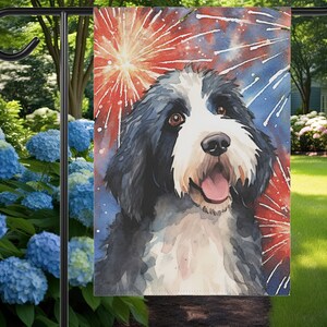 May include: A black and white dog with a happy expression is featured on a garden flag. The dog is surrounded by red, white, and blue fireworks.
