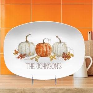 May include: White oval serving platter with a watercolor design of three pumpkins and fall leaves. The pumpkins are white and orange. The text "THE JOHNSON'S" is printed below the pumpkins. The platter is displayed on a wooden surface.