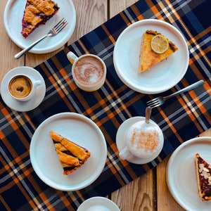 May include: Overhead shot of a wooden table set with white plates, slices of pie, and cups of coffee and cappuccino. A blue, orange, and white plaid tablecloth runs down the center. Silver forks are placed next to the pie slices.