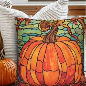 May include: Decorative square pillow featuring a stained-glass style pumpkin design in shades of orange, yellow, and brown. The pumpkin is set against a backdrop of teal and blue. A real pumpkin is next to the pillow.