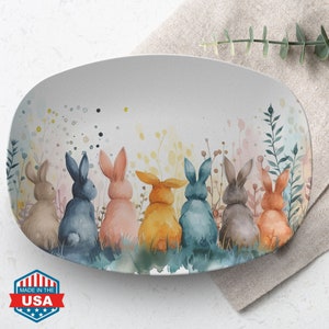 May include: A white oval serving platter with a watercolor design of seven bunnies sitting in a field of flowers. The bunnies are facing away from the viewer and are in various shades of blue, pink, brown, and yellow.