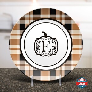 May include: A decorative plate with a pumpkin design and the letter "E". The plate features a brown, black, and white plaid pattern around the rim. The center of the plate is white with a black pumpkin design and the letter "E".