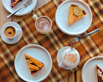May include: A plaid table runner with a brown, orange, and yellow plaid pattern. There are six white plates with various desserts and coffee drinks on top of the runner.