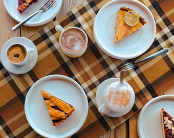 May include: Overhead shot of a wooden table with white plates holding slices of pie and coffee cups. The pies have fruit fillings and a lemon slice. The coffee cups contain espresso, cappuccino, and hot chocolate. A brown and tan plaid table runner is in the center.