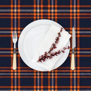 May include: A white dinner plate with a folded white napkin and a sprig of red berries on a blue and orange plaid tablecloth. A white dinner plate and a fork are also on the table.