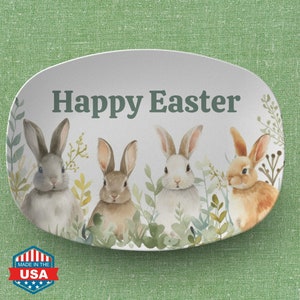 May include: Oval-shaped serving platter with a watercolor design featuring four bunnies and the text "Happy Easter." The bunnies are in shades of gray, brown, and white, surrounded by green foliage. The platter has a white background and is marked "Made in the USA."