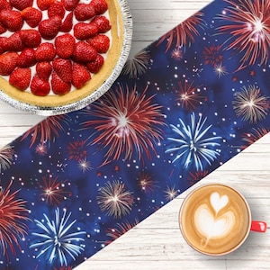 May include: A blue table runner with a red, white, and blue fireworks pattern. The runner is on a white wooden surface with a dessert and a cup of coffee.